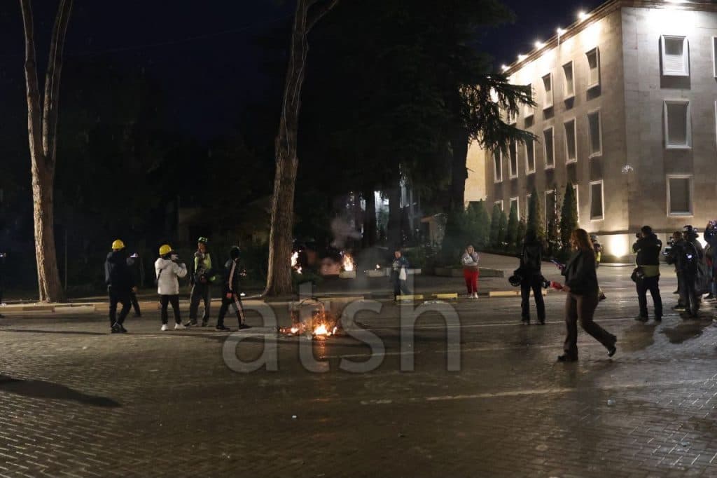 Small fires on the boulevard near the illuminated Prime Minister's Office building, with protesters and journalists at night