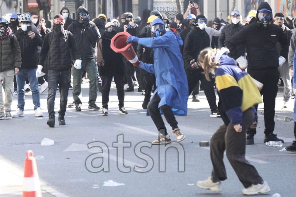 Protesters wearing masks and goggles in the street during the protest in Tirana; one holds a traffic cone