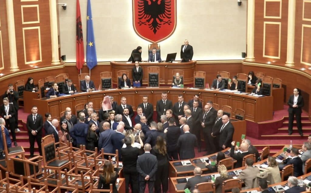 Deputetë të opozitës dhe të shumicës të grumbulluar para foltores në sallën e Kuvendit, me karrige të përmbysura në plan të parë